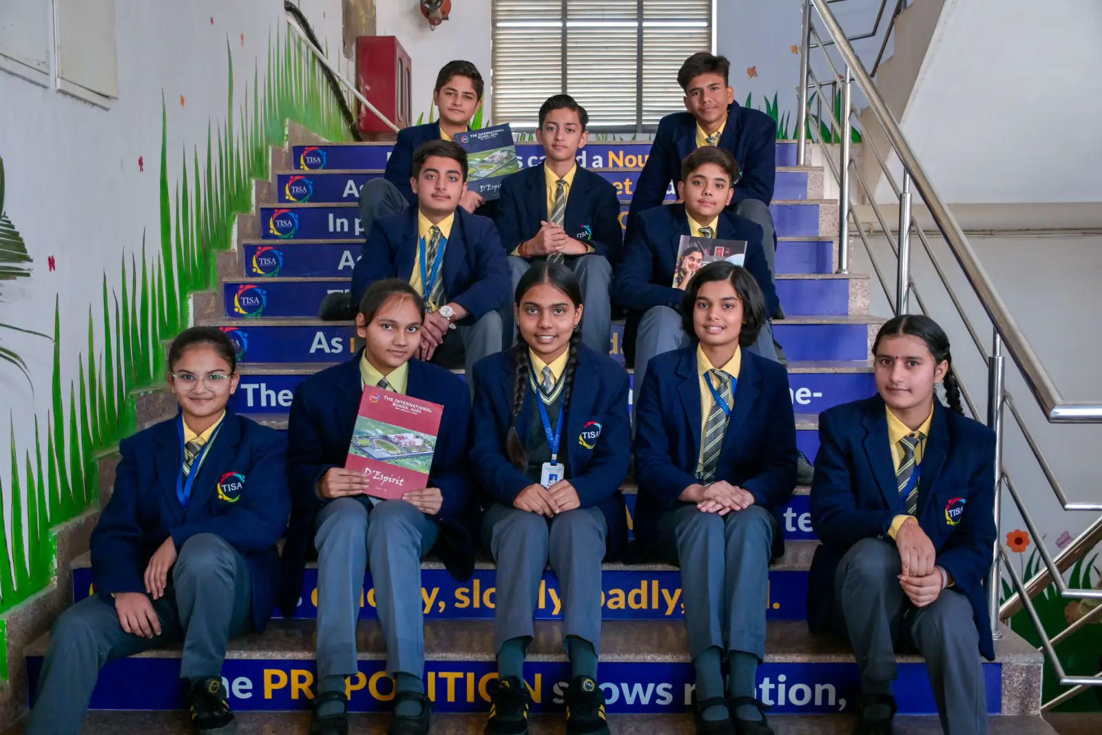 A group of tisa students in formal school uniforms sitting on decorated stairs inside the school, holding notebooks and smiling confidently for a group photo.