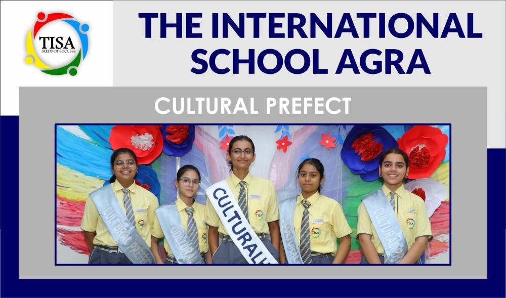 Group photo of cultural prefects at the international school agra. Five students stand in front of a vibrant, artistic backdrop decorated with large red, blue, and white paper flowers. They are dressed in yellow school shirts and grey trousers. Three of the students wear light blue sashes labeled "cultural prefect" while the others are formally dressed as well, symbolizing leadership. Above the image is the tisa logo with the text "the international school agra" and "cultural prefect" written in bold. The design reflects school spirit, leadership, and celebration of cultural excellence.