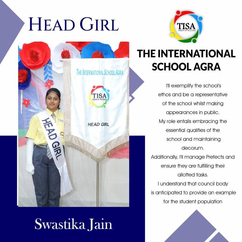 Portrait of swastika jain, head girl of the international school agra, wearing a yellow shirt, grey trousers, and a white sash labeled "head girl". She stands beside a banner with the school logo and the title "head girl", with a backdrop of decorative red, white, and blue paper flowers. The text beside her outlines her commitment to representing the school’s values, maintaining decorum, managing prefects, and setting an example for the student body. The school logo and name are prominently displayed at the top.