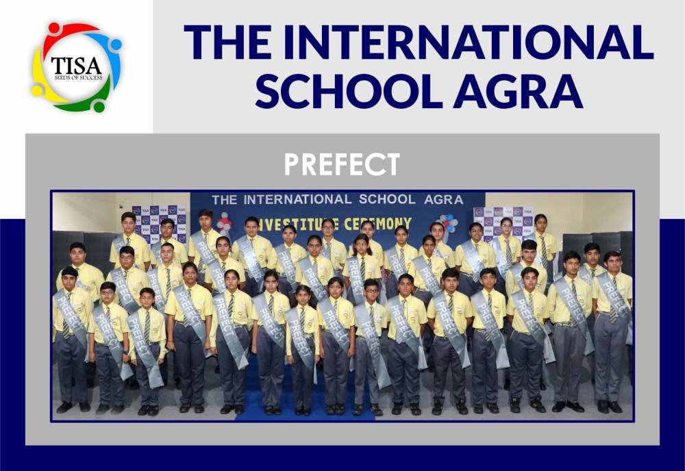 Group photo of prefects at the international school agra during the investiture ceremony. Students are standing in rows, wearing yellow shirts, grey trousers, and grey sashes labeled "prefect". A banner in the background reads "the international school agra - investiture ceremony". The school logo with the tagline "seeds of success" is displayed in the top left corner.