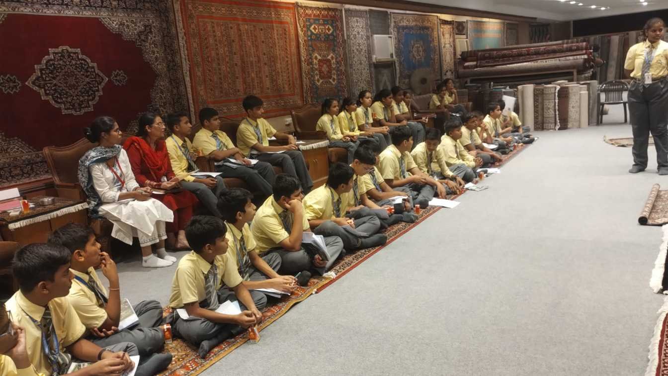 A group of school students in yellow uniforms sit on rugs inside a carpet showroom or gallery, attentively listening to a presentation.